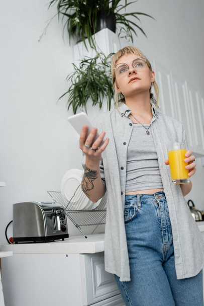 wytatuowana młoda kobieta w okularach, z pierścieniem na palcu trzymającym szklankę soku pomarańczowego i smartfona podczas pisania SMS-ów i stania w rozmytej kuchni z tosterem i roślinami wewnątrz w nowoczesnym apartamencie  - Zdjęcie, obraz