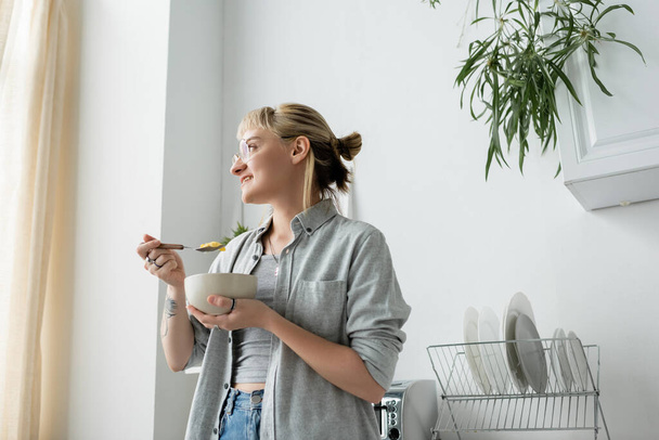 getatoeëerde jonge vrouw met pony en bril glimlachend terwijl het vasthouden van kom met cornflakes en lepel tijdens het ontbijt en wegkijken in de buurt van groene planten en schone gerechten in de witte keuken  - Foto, afbeelding