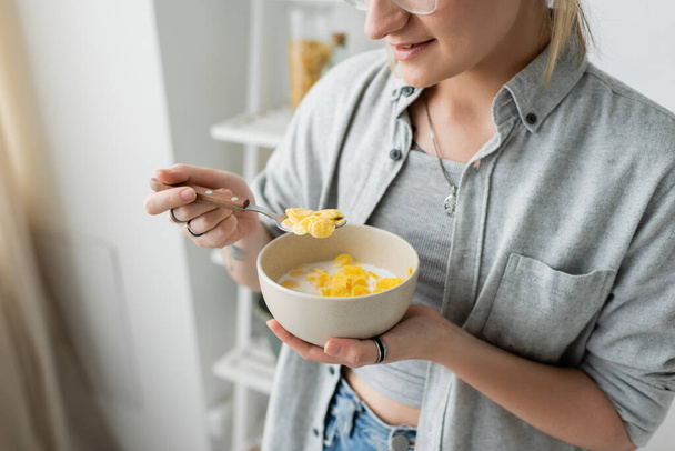 cropped view of tattooed young woman smiling while holding bowl with cornflakes and spoon while having breakfast next to blurred white rack in modern kitchen. copy space, apartment, morning energy  - Photo, Image