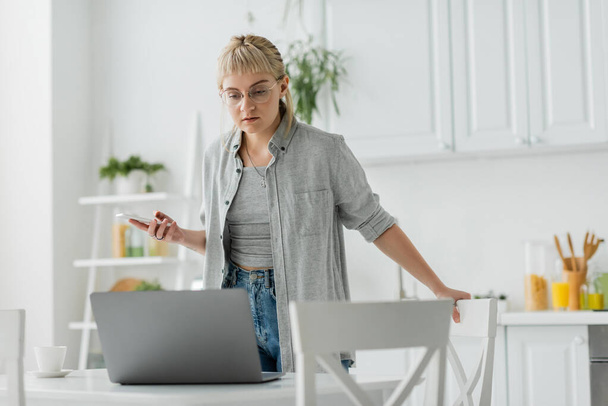 Kısa saçlı, dövmeli ve kâküllü genç bir kadın akıllı telefon kullanırken bir fincan kahve bardağının yanında duruyor ve modern apartmandaki sandalyelerin etrafındaki bulanık laptopa bakıyor.  - Fotoğraf, Görsel