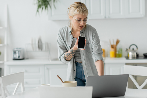 young woman with bangs and tattoo on hand recording voice message on smartphone while standing in eyeglasses, bowl with cornflakes, cup of coffee and laptop on table in modern apartment, freelancer  - Photo, Image