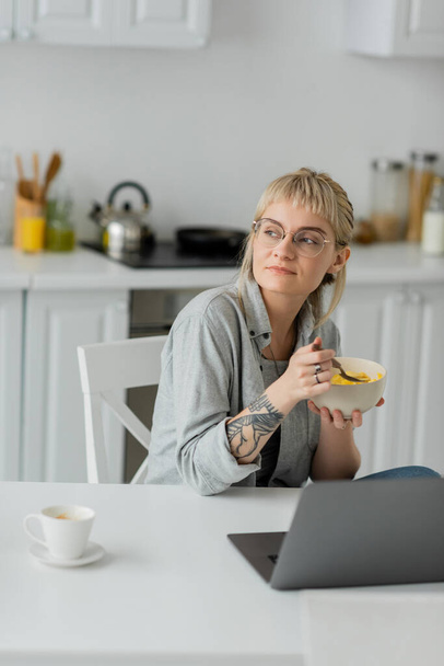 marzycielska młoda kobieta z grzywką, krótkimi włosami i tatuażem na ręku jedząca płatki kukurydziane na śniadanie podczas korzystania z laptopa przy filiżance kawy ze spodkiem na stole w nowoczesnej kuchni, freelancer, praca w domu  - Zdjęcie, obraz