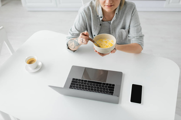 abgeschnittene Ansicht einer jungen Frau mit Tätowierung auf der Hand, die Cornflakes zum Frühstück isst, während sie Laptop in der Nähe von Smartphone mit leerem Bildschirm und Tasse Kaffee auf dem Tisch in der modernen Küche benutzt, freiberuflich  - Foto, Bild