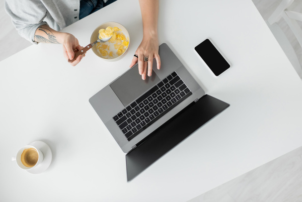 vista superior de mujer joven con tatuaje en la mano comer copos de maíz para el desayuno mientras se utiliza el ordenador portátil cerca de teléfono inteligente con pantalla en blanco y taza de café en la mesa en la cocina moderna, freelancer, tiro recortado - Foto, Imagen