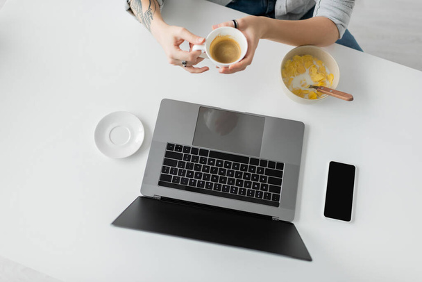 верхний вид женщины с татуировкой на руке держа чашку кофе возле миски с кукурузными хлопьями во время завтрака при использовании ноутбука рядом со смартфоном с чистым экраном в современной кухне, фрилансер, обрезанный  - Фото, изображение