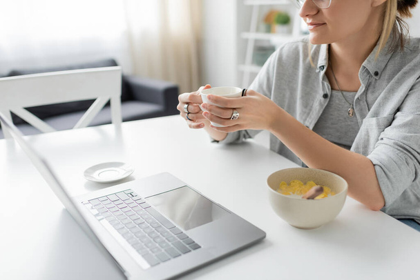 recortado tiro de mujer joven sosteniendo taza de café cerca de tazón con copos de maíz durante el desayuno, mientras que el uso de la computadora portátil cerca de platillo blanco en la mesa en la cocina moderna, freelancer, trabajo desde casa  - Foto, Imagen