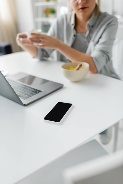 abgeschnittene Aufnahme einer verschwommenen Frau, die während des Frühstücks eine Tasse Kaffee in der Nähe einer Schüssel mit Cornflakes hält, während sie Laptop in der Nähe eines Smartphones mit leerem Bildschirm in der modernen Küche benutzt, Freelancer  - Foto, Bild