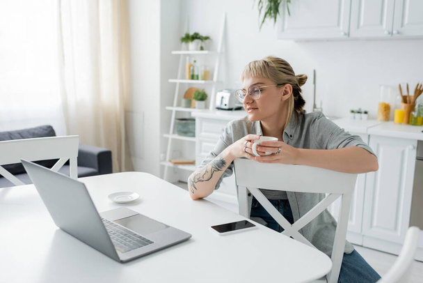 mujer joven con tatuaje en la mano y flequillo sosteniendo taza de café y mirando a la computadora portátil cerca de teléfono inteligente y platillo en la mesa blanca alrededor de sillas en la cocina moderna, freelancer, estilo de vida remoto  - Foto, imagen