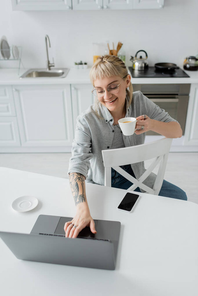 над головой вид счастливой молодой женщины с татуировкой на руке и челкой с чашкой кофе и глядя на ноутбук рядом со смартфоном и блюдцем на белом столе вокруг стульев в современной кухне, удаленный образ жизни - Фото, изображение