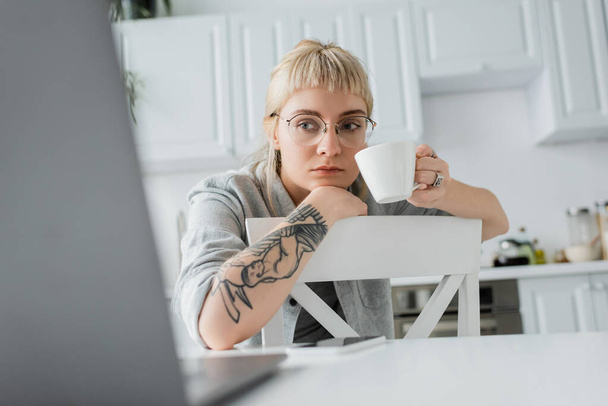 skupiona młoda kobieta z tatuażem na dłoni i grzywką trzymająca filiżankę kawy i patrząca na niewyraźny laptop w pobliżu smartfona na białym stole w nowoczesnej kuchni, freelancer, zdalny styl życia  - Zdjęcie, obraz