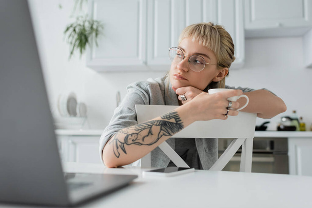 marzycielska młoda kobieta z tatuażem na dłoni i grzywką trzymająca filiżankę kawy i odwracająca wzrok w pobliżu zamazanego laptopa i smartfona na białym stole w nowoczesnej kuchni, freelancer, zdalny styl życia  - Zdjęcie, obraz