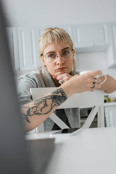 Набридлива молода жінка в окулярах з татуюванням на руках і банками, що тримають чашку кави, і дивиться на ноутбук біля смартфона на білому столі на сучасній кухні, фрілансер, віддалений спосіб життя.  - Фото, зображення