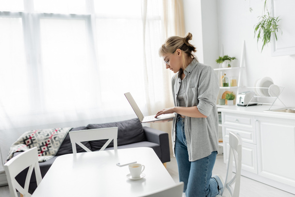 fiatal nő szemüveg rövid haj és frufru gazdaság laptop közelében csésze kávé és okostelefon fehér asztal körül székek fehér és modern konyha mellett kanapé, szabadúszó, távoli életmód - Fotó, kép