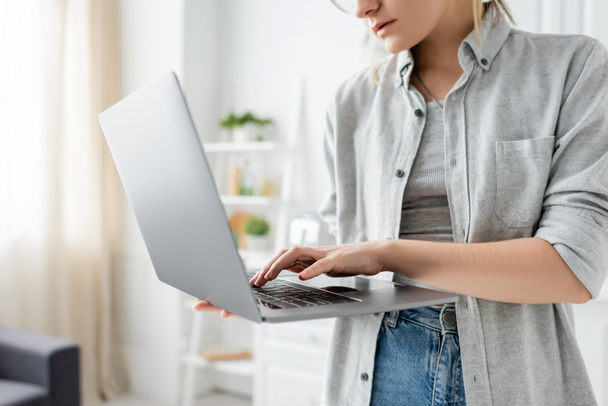 ostříhaný pohled na zaostřenou mladou ženu v šedé košili držící a používající notebook v bílé a moderní kuchyni, rozmazané pozadí, vzdálený životní styl, na volné noze  - Fotografie, Obrázek