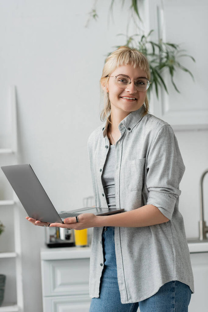 ευτυχισμένη νεαρή γυναίκα με γυαλιά ηλίου, κοντά μαλλιά και αφέλειες κρατώντας και χρησιμοποιώντας φορητό υπολογιστή, ενώ κοιτάζοντας κάμερα και εργάζονται από το σπίτι στη σύγχρονη κουζίνα, θολή φόντο, απομακρυσμένο τρόπο ζωής, ελεύθερος επαγγελματίας - Φωτογραφία, εικόνα