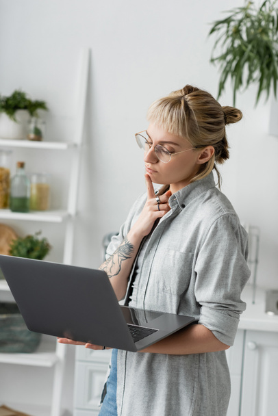 pensive young woman in eyeglasses, with tattoo and bangs using laptop while working from home in white and modern kitchen, blurred background, remote lifestyle, freelancer, hand near chin - Photo, Image