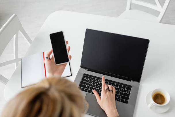 widok z góry rozmyty kobieta gospodarstwa smartfon z pustym ekranem w pobliżu laptopa, notebook z piórem i filiżanka kawy z talerzykiem na białym stole podczas pracy w domu, freelancer, nowoczesne miejsce pracy  - Zdjęcie, obraz