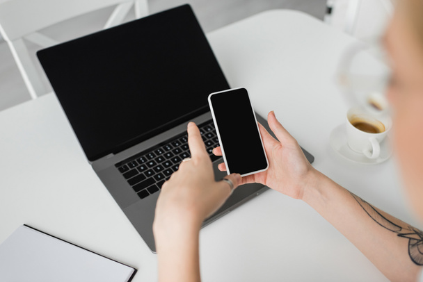 widok z góry wytatuowany kobieta gospodarstwa smartfon z pustym ekranem w pobliżu laptopa, notebook, i filiżanka kawy ze spodkiem na białym stole podczas pracy w domu, freelancer, nowoczesne miejsce pracy  - Zdjęcie, obraz