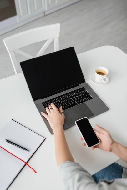widok z góry kobieta gospodarstwa smartfon z pustym ekranem i za pomocą laptopa w pobliżu notebooka z długopisem, i filiżanka kawy ze spodkiem na białym stole podczas pracy w domu, freelancer, nowoczesne miejsce pracy  - Zdjęcie, obraz