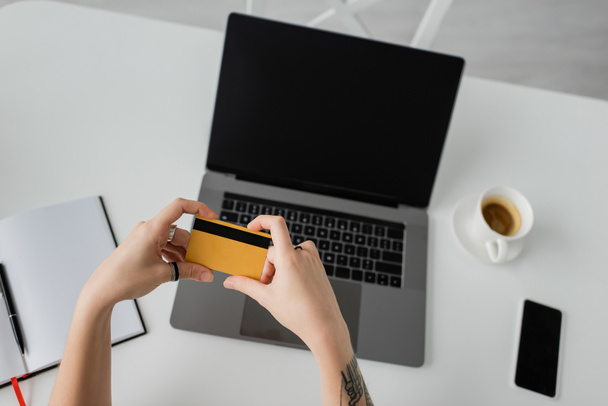 top view tetovált nő gazdaság hitelkártya közelében laptop üres képernyő, okostelefon, notebook toll, és egy csésze kávé csészealj fehér asztalon, modern munkaterület, munka otthonról  - Fotó, kép