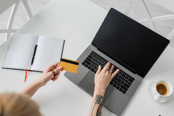 widok z góry wytatuowanej młodej kobiety trzymającej kartę kredytową i korzystającej z laptopa z pustym ekranem w pobliżu notebooka z długopisem, filiżanka kawy ze spodkiem na białym stole, nowoczesne miejsce do pracy, praca w domu  - Zdjęcie, obraz