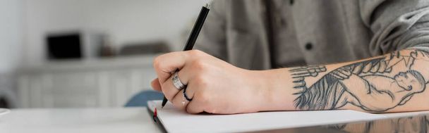 kivágott felvétel fiatal nő tetoválás kézírás notebook, jegyzetelés, amelynek ihletet, miközben a kezében toll mellett laptop fehér asztalon, elmosódott előtér, munka otthonról, banner  - Fotó, kép