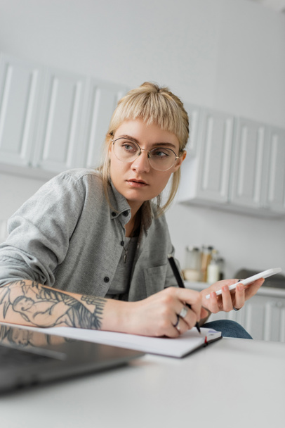 mladá žena s tetováním na ruce psaní do notebooku, psaní poznámek, držení smartphonu a pera u notebooku na bílém stole, rozmazané popředí, práce z domova  - Fotografie, Obrázek