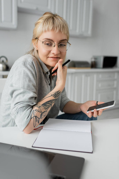 szczęśliwa młoda kobieta z tatuażem na dłoni i grzywką trzymając smartfona z pustym ekranem i długopisem w pobliżu notebooka i laptopa na białym stole, rozmyty pierwszy plan, praca w domu  - Zdjęcie, obraz
