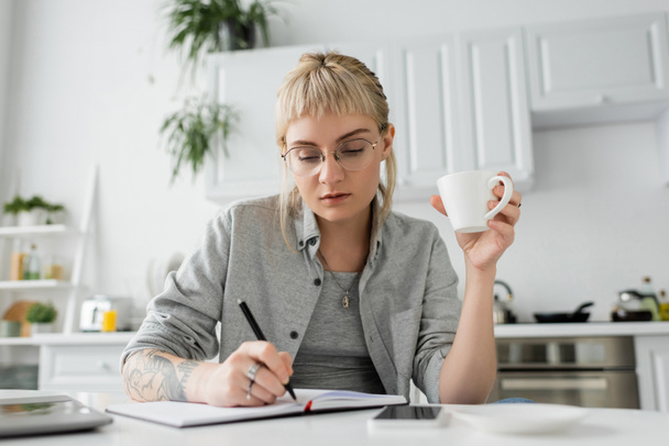 молодая женщина с татуировкой на руке и челкой, держа чашку кофе возле ноутбука, смартфона и ноутбука на белом столе, размытый передний план, работа из дома, делать заметки  - Фото, изображение