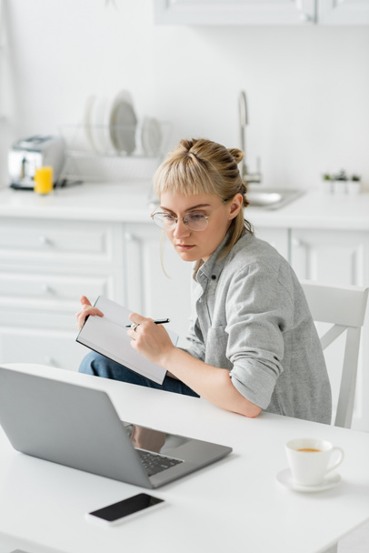 молодая женщина с татуировкой на руке и челкой, держащая ноутбук, делая заметки возле смартфона и ноутбука на белом столе, размытый фон, работа из дома  - Фото, изображение