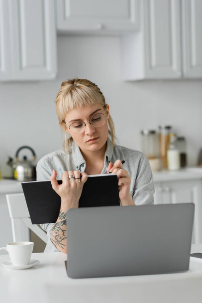 giovane donna in occhiali con tatuaggio sulla mano e frangia che tiene il taccuino, prendendo appunti, seduta vicino al computer portatile e alla tazza di caffè sul tavolo bianco, sfondo sfocato, lavoro da casa  - Foto, immagini