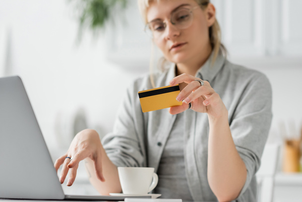 junge Frau mit Brille und tätowierter Kreditkarte in der Hand, neben Laptop und Kaffeetasse auf weißem Tisch sitzend, verschwommener Hintergrund, Arbeit von zu Hause aus, Online-Transaktionen, Technologie  - Foto, Bild