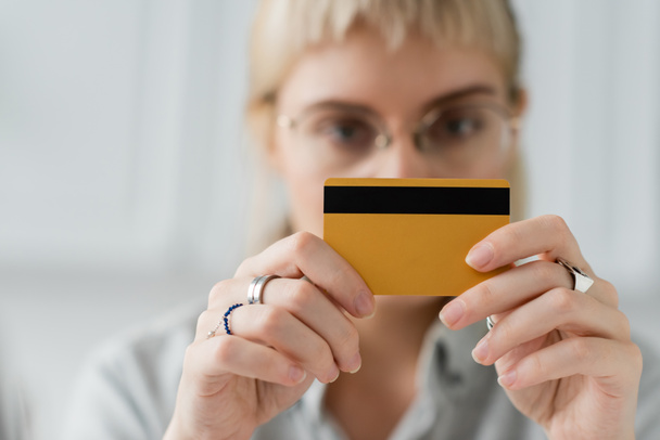 размытая сцена молодой женщины в очках с кольцами на пальцах, держащих кредитную карту в руках и смотрящей в камеру дома с размытым фоном, копировальным пространством  - Фото, изображение