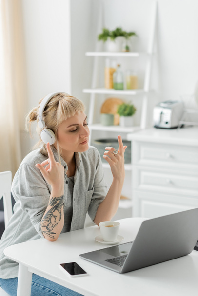 młoda kobieta z grzywką i tatuażem na dłoni siedzi w słuchawkach bezprzewodowych i gestykulując przy laptopie, filiżance kawy i niewyraźnym smartfonie z nieostrym ekranem na stole, freelance, praca w domu - Zdjęcie, obraz