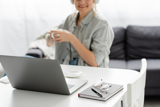очки и ручка на ноутбуке рядом с ноутбуком и веселый фрилансер улыбается, держа чашку кофе на размытом фоне дома, работа из дома, современное рабочее пространство  - Фото, изображение