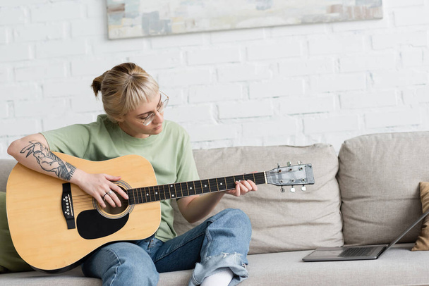 mujer joven en gafas con flequillo y tatuaje sosteniendo la guitarra acústica y aprender a jugar mientras busca video tutorial en el ordenador portátil y sentado en cómodo sofá en la sala de estar moderna en casa - Foto, Imagen