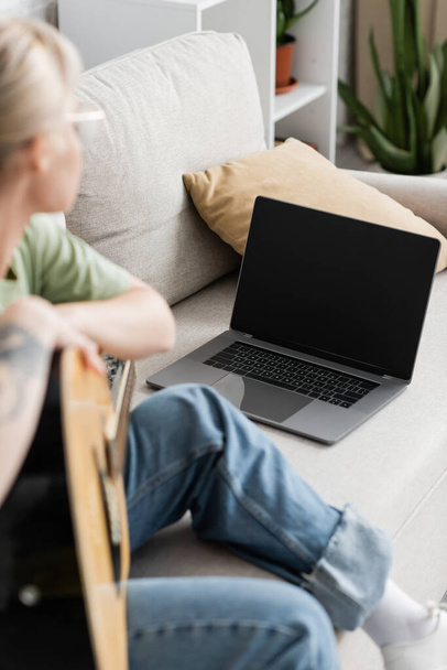 jonge vrouw in bril met akoestische gitaar en leren spelen tijdens het kijken video tutorial op laptop met leeg scherm en zitten op een comfortabele bank in de woonkamer, gitaar lessen - Foto, afbeelding