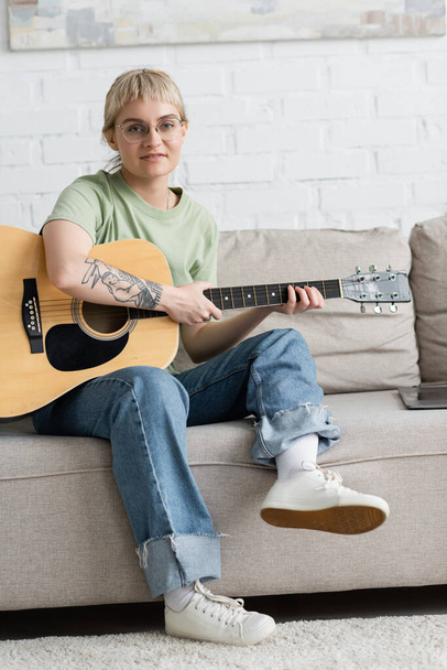 fiatal nő szemüveg frufru és tetoválás gazdaság akusztikus gitár és megtanulják, hogyan kell játszani a laptop közelében, és ül kényelmes kanapén, nézi a kamera a modern nappaliban otthon  - Fotó, kép