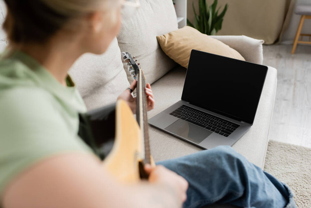 Bulanık genç bir kadın elinde akustik gitar tutarken ve bilgisayar başında boş ekranla video dersi alırken ve oturma odasında rahat bir koltukta otururken çalmayı öğrenirken. - Fotoğraf, Görsel