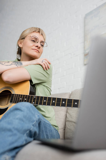 довольная молодая женщина в очках с челкой и татуировкой, держащая акустическую гитару и глядя видео-учебник на ноутбуке, сидя на удобном диване в современной гостиной дома, цифровые ресурсы  - Фото, изображение