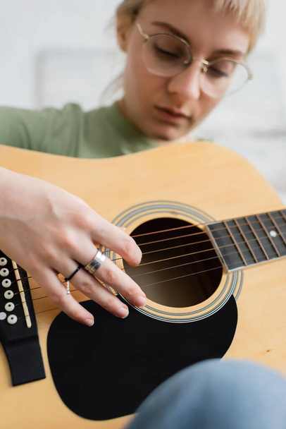 talanted jovem mulher em óculos com franja, anéis em dedos tocando guitarra acústica e sentado na sala de estar moderna, aprendendo música, desenvolvimento de habilidades, entusiasta da música  - Foto, Imagem