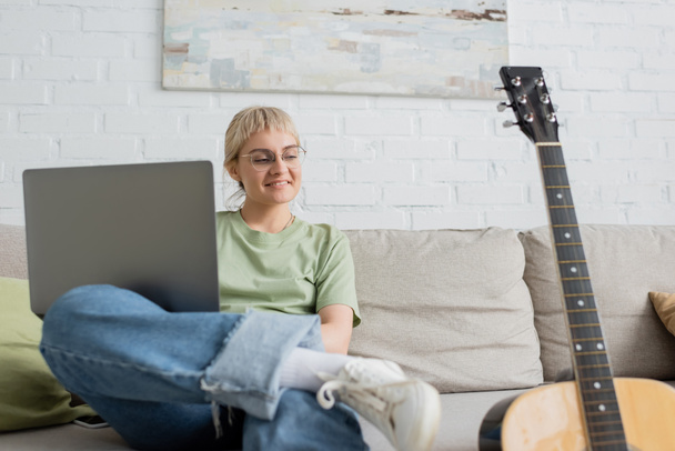 happy young woman with blonde and short hair, bangs and eyeglasses using laptop while sitting on comfortable couch and looking at guitar in modern living room with painting on wall  - Фото, изображение