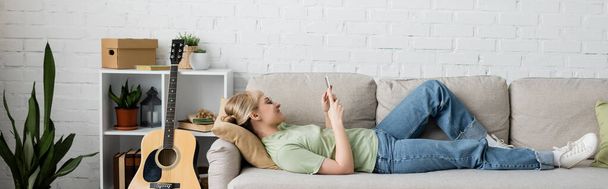 side view of happy woman with blonde and short hair, bangs and eyeglasses using smartphone while resting on comfortable couch near guitar in modern living room with rack and plants, banner - Photo, Image