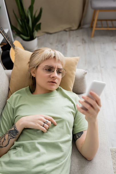 Overhead-Ansicht einer jungen Frau mit Tätowierung, blonden und kurzen Haaren, Pony und Brille mittels Smartphone, während sie auf einer bequemen Couch neben der Gitarre im modernen Wohnzimmer ruht, verschwommener Hintergrund - Foto, Bild