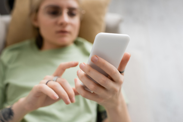 overhead view of young woman with tattoo, blonde and short hair, bangs and eyeglasses using smartphone while resting on comfortable couch in modern living room, blurred background - Photo, Image