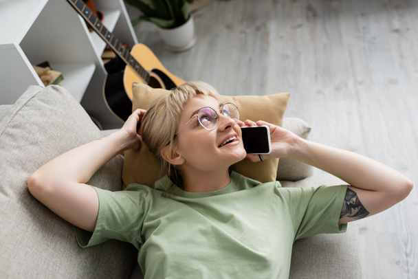 над головой вид счастливой молодой женщины с короткими и светлыми волосами, челкой и очками, разговаривающей на смартфоне, отдыхающей на удобном диване рядом с гитарой в современной гостиной  - Фото, изображение