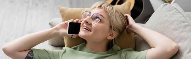 overhead view of happy young woman with blonde and short hair, bangs and eyeglasses talking on smartphone while resting on comfortable couch near guitar in modern living room, banner  - Photo, Image