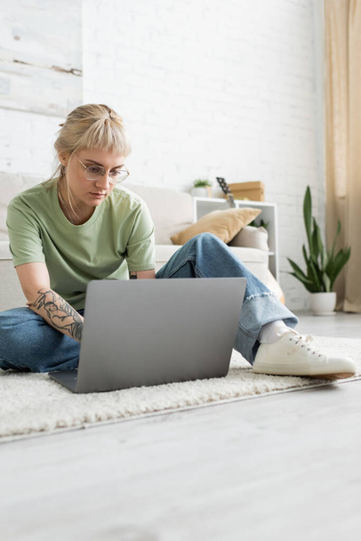 wytatuowana kobieta z blond włosami, grzywką i okularami za pomocą laptopa, siedząca na dywanie w pobliżu wygodnej kanapy, rozmyta roślina i stojak w nowoczesnym salonie z malowaniem na ścianie  - Zdjęcie, obraz
