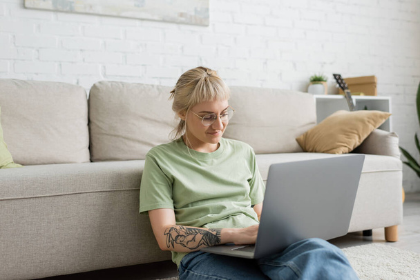 Tätowierte Frau mit blonden und kurzen Haaren, Pony und Brille tippt auf Laptop, während sie auf Teppich in der Nähe einer bequemen Couch im modernen Wohnzimmer mit Wandmalerei sitzt  - Foto, Bild