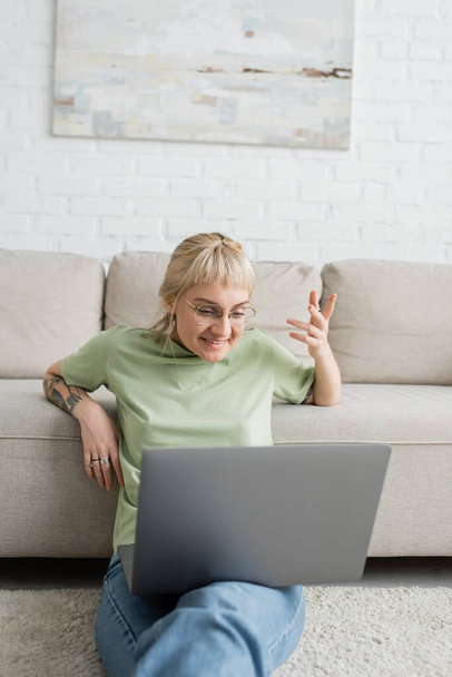wytatuowana kobieta z blond i krótkimi włosami, grzywką i okularami uśmiechnięta podczas rozmowy wideo na laptopie siedząc na dywanie przy wygodnej kanapie w nowoczesnym salonie z malowaniem na ścianie  - Zdjęcie, obraz